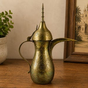 May include: A brass coffee pot with a tall, pointed lid, a curved handle, and a long, slender spout. The body of the vessel features intricate floral engravings, while the metal surface displays a weathered, aged patina with dark highlights.