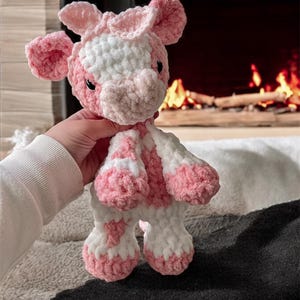 May include: A pink and white crocheted cow plush toy. The toy has a white face with pink spots and pink ears. The toy is being held by a hand.