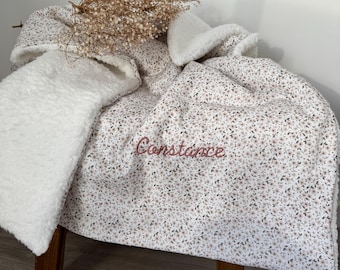 May include: A floral-patterned blanket with a white, fluffy interior is draped over a small wooden stool. The blanket features a delicate pattern of small flowers in shades of brown and pink. The name "Constance" is embroidered in a cursive font.