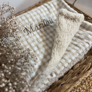 May include: A folded blanket featuring a beige and white gingham pattern on one side and a cream-colored sherpa fleece texture on the other. The name Marilou is embroidered in brown thread on the gingham side of the fabric.