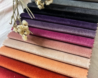 May include: A collection of velvet fabric swatches in various colors, including shades of red, orange, pink, and purple. The swatches are arranged in a layered display, showcasing the texture and color variations. Dried flowers are placed on top of the swatches.