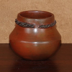 Puede incluir: Una pequeña maceta redonda de terracota con una banda marrón oscura alrededor de la parte superior. La maceta está sobre una superficie de madera marrón oscura.
