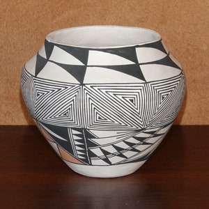 Puede incluir: Una olla de cerámica blanca con diseños geométricos negros y naranja. La olla está decorada con un patrón tradicional del suroeste.