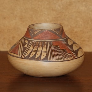 Puede incluir: Un pequeño jarrón redondo de cerámica marrón con diseños geométricos en rojo y negro. El jarrón está sobre una superficie de madera.