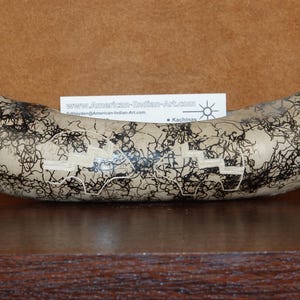 May include: A curved, elongated ceramic vessel with a light beige surface featuring intricate black horsehair-style markings and carved geometric step patterns. A white rectangular card with the text www.American-Indian-Art.com is visible.