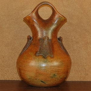 May include: A brown ceramic Native American pottery vase with a unique design. The vase has a wide base and a narrow neck with a handle. The vase is decorated with a geometric pattern.