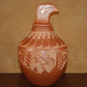 Può includere: Un vaso in terracotta fatto a mano con un design unico. Il vaso ha un coperchio con una testa di uccello scolpita e un motivo geometrico.