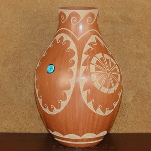 Può includere: Un vaso in terracotta con un design geometrico e un'intarsio di pietra turchese. Il vaso è decorato con linee e spirali bianche.