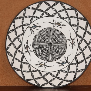 Puede incluir: Un plato de cerámica blanco con un diseño geométrico en blanco y negro. El plato presenta un patrón repetitivo de círculos y cuadrados. Hay seis lagartos negros con marcas blancas en el plato.