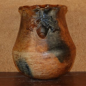 May include: A brown and black ceramic pot with a unique design. The pot has a wide base and a narrow neck.