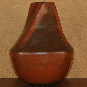 May include: A dark brown and red ceramic vase with a unique, angular shape. The vase has a wide base and a narrow neck.