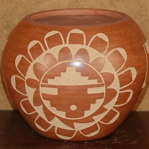 May include: A terracotta pottery vase with a carved design of a sun face with a geometric pattern. The vase is decorated with a white slip.