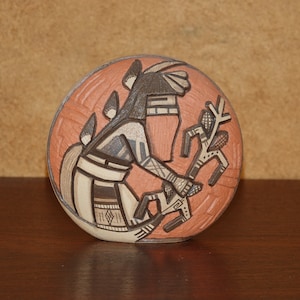 Puede incluir: Un azulejo de cerámica redondo pintado a mano con un diseño nativo americano. El azulejo presenta una figura estilizada en negro, blanco y marrón, sosteniendo una rama con hojas. El fondo es de color naranja rojizo.