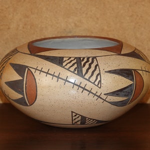 May include: A tan ceramic bowl with a geometric design in brown and black. The bowl has a rounded shape and a smooth surface.