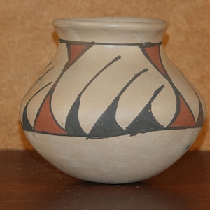 May include: A small, rounded ceramic pot with a matte cream-colored base. The upper section features a repeating geometric pattern consisting of alternating black and terracotta-colored triangles and diagonal lines encircling the vessel.