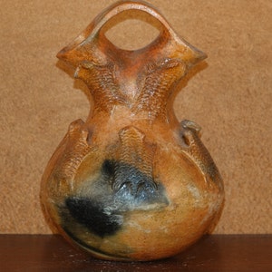 May include: A brown and black ceramic Native American pottery vase with a unique design. The vase has a wide base and a narrow neck with a handle.