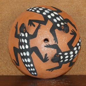 Può includere: Un oggetto rotondo in ceramica color terracotta con tre lucertole nere stilizzate con punti bianchi dipinti sulla superficie. Le lucertole sono disposte attorno a un foro centrale.