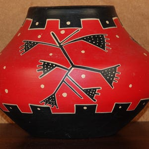 May include: A rounded ceramic pot featuring a vibrant red body with a matte finish. The vessel is decorated with black geometric patterns, including a central stylized plant motif and jagged borders at the top and bottom, accented by small white dots.