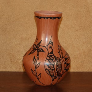 Op de afbeelding: Een terracotta vaas met een geometrisch ontwerp in zwart en wit van een vogel en een bloem. De vaas heeft een smalle hals en een rond lichaam.