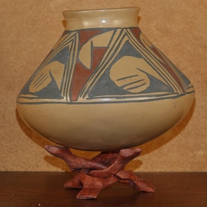 May include: A rounded ceramic pot with a wide body and a narrow neck, featuring a matte tan base color decorated with geometric patterns in dark gray and terracotta red. The vessel is supported by a dark reddish-brown wooden tripod stand.