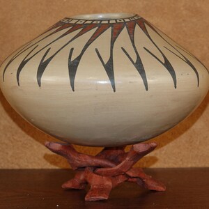May include: A round, beige ceramic vessel with a wide, tapered body and a narrow opening. The top section features a decorative pattern of dark brown, pointed, radiating shapes and a border of small squares. It rests on a reddish-brown wooden stand.