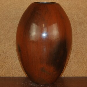 May include: A tall, oval-shaped vase with a glossy, reddish-brown finish. The ceramic vase has dark brown accents and a dark rim. The vase is displayed on a dark brown surface against a tan background.