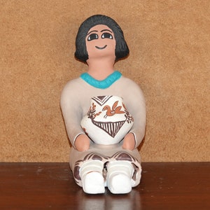 May include: A hand-painted ceramic figurine of a woman sitting with a pot in her lap. The woman has black hair, brown eyes, and a light brown complexion. She is wearing a white dress with a turquoise necklace. The pot is white with brown and orange designs.