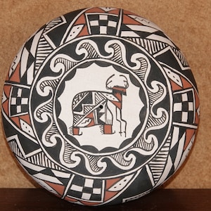 Puede incluir: Un cuenco de cerámica negro, blanco y rojo con un diseño geométrico. El centro del cuenco presenta una figura de animal estilizada.