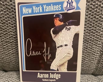 May include: A baseball card featuring Aaron Judge of the New York Yankees. The card has a blue and white border with the team logo and the words "New York Yankees" at the top. Judge is shown in his pinstripe uniform, swinging a bat. The card is signed.