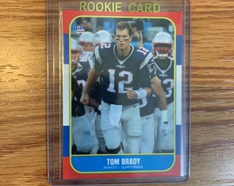 Puede incluir: Una tarjeta de novato de Tom Brady, un jugador de fútbol americano de los New England Patriots. La tarjeta es azul, roja y blanca con letras doradas. La tarjeta presenta una foto de Brady con su uniforme, corriendo con el balón. El texto en la tarjeta dice "Rookie Card", "RC", "Tom Brady", "Patriots" y "Quarterback".