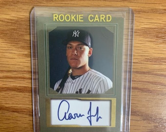May include: A green and gold rookie card with a photo of a baseball player wearing a New York Yankees uniform. The player is holding a baseball bat and has a signature that reads "Aaron Judge".