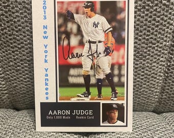 May include: A baseball card featuring Aaron Judge, a New York Yankees player. The card is from 2013 and is a rookie card. The card is white with blue and red accents. The card has a picture of Aaron Judge in his Yankees uniform, and the text "2013 New York Yankees" on the left side of the card. The card also has the text "Aaron Judge" and "Rookie Card" on the bottom of the card.