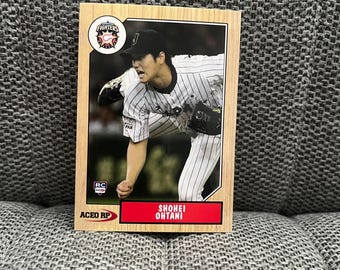 May include: A baseball trading card featuring Shohei Ohtani in a Hokkaido Nippon-Ham Fighters uniform. The card has a wooden frame with the team logo and player's name. The background shows Ohtani pitching.