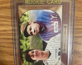 May include: A baseball rookie card featuring a photo of two baseball players. The card has a gold border and a wooden background. The text "ROOKIE CARD" is printed in gold at the top of the card. The player on the left is wearing a blue and white baseball uniform with the name "RUTH" printed on the back. The player on the right is wearing a white and black baseball uniform with the name "OTANI" printed on the back.
