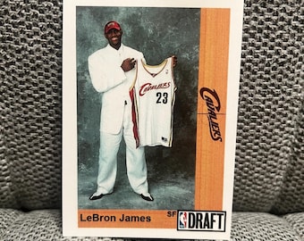 Puede incluir: Una tarjeta de baloncesto con LeBron James con un traje blanco sosteniendo una camiseta de los Cleveland Cavaliers. La camiseta es blanca con detalles granates y dorados, y el número 23 es visible. La tarjeta incluye el texto "LeBron James" y "DRAFT".