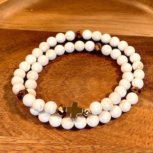 May include: A white and brown beaded bracelet with a gold cross charm. The bracelet is made of small, round beads and has a simple, elegant design.