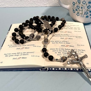 May include: A black rosary with a silver crucifix and a silver charm. The rosary is resting on an open book with text about prayer. The book is open to pages 42 and 43.