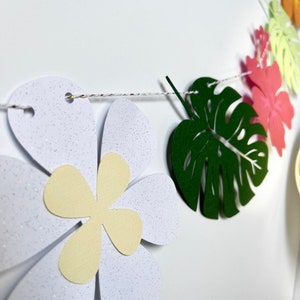 Pode incluir: Uma guirlanda de papel com tema tropical com flores brancas, folhas verdes e flores cor-de-rosa. A guirlanda &eacute; enfiada com um barbante branco.