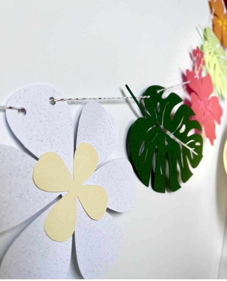 Pode incluir: Uma guirlanda de papel com tema tropical com flores brancas, folhas verdes e flores cor-de-rosa. A guirlanda &eacute; enfiada com um barbante branco.
