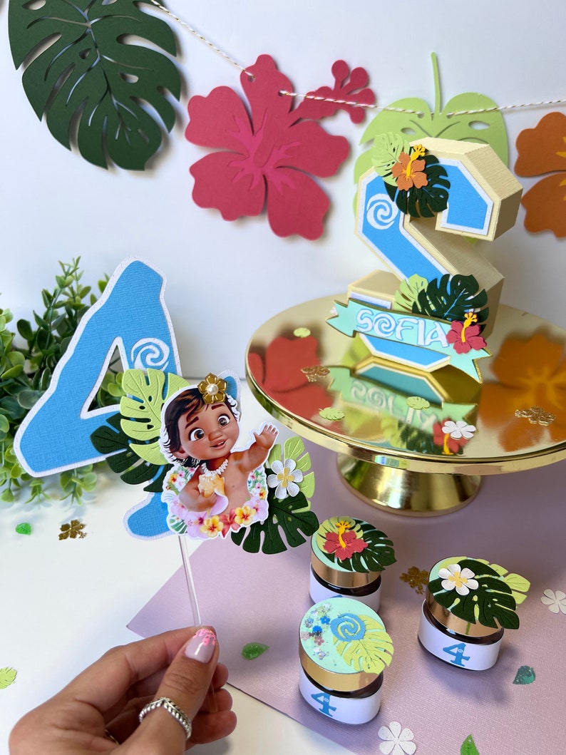 Pode incluir: Um topper de bolo azul com o n&uacute;mero quatro, um personagem de Moana, folhas verdes e um design de espiral. H&aacute; tamb&eacute;m pequenos frascos com o n&uacute;mero quatro e decora&ccedil;&otilde;es tropicais.