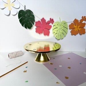 Pode incluir: Um suporte de bolo dourado sobre um fundo branco. Uma corda de folhas e flores tropicais de papel pendura sobre o suporte. As folhas s&atilde;o verdes, amarelas e laranja. As flores s&atilde;o brancas, cor-de-rosa e laranja.