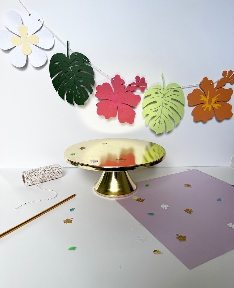 Pode incluir: Um suporte de bolo dourado sobre um fundo branco. Uma corda de folhas e flores tropicais de papel pendura sobre o suporte. As folhas s&atilde;o verdes, amarelas e laranja. As flores s&atilde;o brancas, cor-de-rosa e laranja.