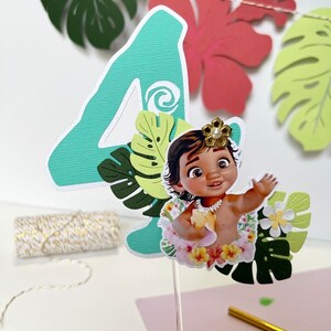 Pode incluir: Um topper de bolo de n&uacute;mero quatro verde com um personagem de Moana no centro. O personagem est&aacute; usando um colar de flores e segurando uma concha. O topper &eacute; cercado por folhas e flores verdes.
