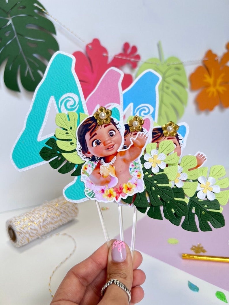 Pode incluir: Tr&ecirc;s toppers de bolo com um personagem de desenho animado do filme Moana. O personagem &eacute; uma beb&ecirc; Moana que usa um colar de flores e segura uma flor. Os toppers s&atilde;o decorados com folhas verdes e flores brancas. Os n&uacute;meros 4 e 1 s&atilde;o vis&iacute;veis no fundo.