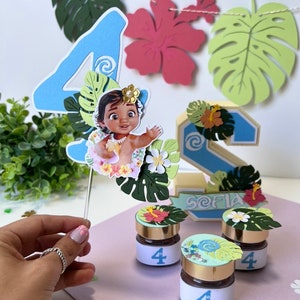 Pode incluir: Um topper de bolo de n&uacute;mero quatro azul com um personagem de Moana, folhas verdes e flores brancas. Tr&ecirc;s pequenos frascos com tampa dourada e um r&oacute;tulo branco com o n&uacute;mero quatro. Os frascos s&atilde;o decorados com folhas verdes e flores. Os frascos est&atilde;o sobre uma superf&iacute;cie rosa. O topper de bolo est&aacute; sobre uma superf&iacute;cie branca.