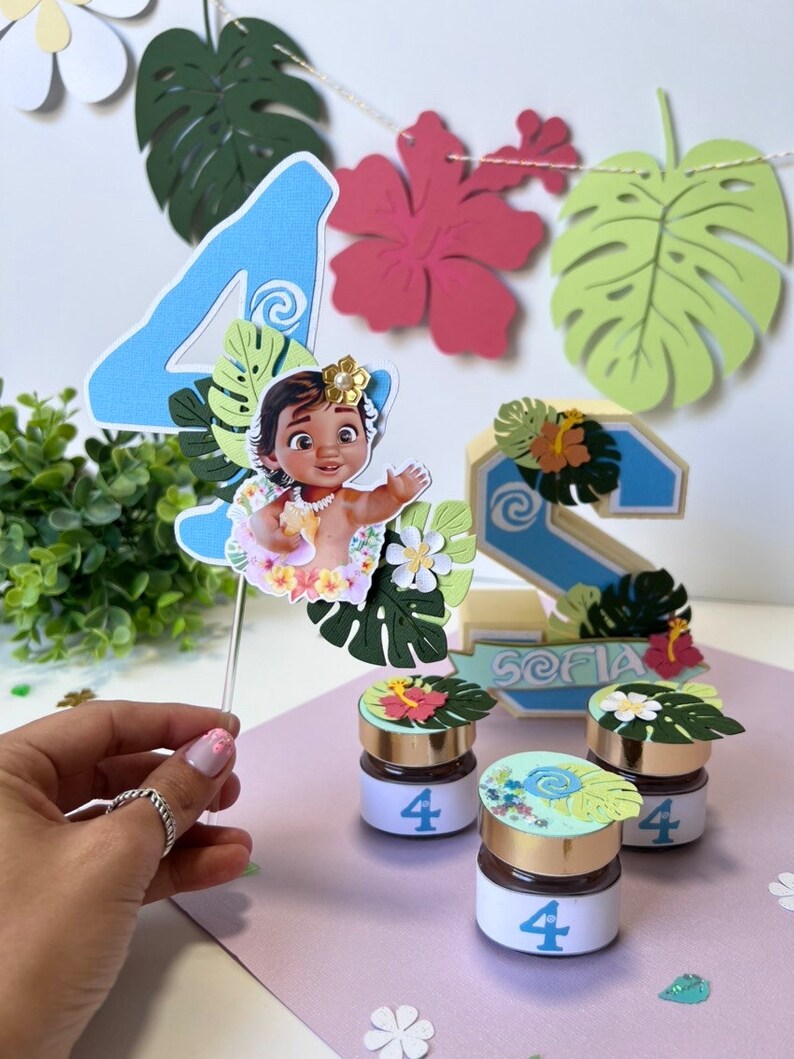 Pode incluir: Um topper de bolo de n&uacute;mero quatro azul com um personagem de Moana, folhas verdes e flores brancas. Tr&ecirc;s pequenos frascos com tampa dourada e um r&oacute;tulo branco com o n&uacute;mero quatro. Os frascos s&atilde;o decorados com folhas verdes e flores. Os frascos est&atilde;o sobre uma superf&iacute;cie rosa. O topper de bolo est&aacute; sobre uma superf&iacute;cie branca.