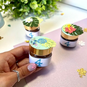 Pode incluir: Tr&ecirc;s pequenos frascos de vidro com tampas douradas e etiquetas brancas com o n&uacute;mero "4". Cada frasco tem uma tampa de papel de tema tropical diferente com flores e folhas.
