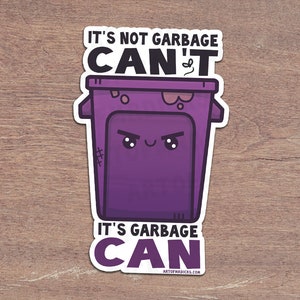 May include: A sticker featuring a purple cartoon trash can with an angry face. The text reads "IT'S NOT GARBAGE CAN'T, IT'S GARBAGE CAN" in black and white. The sticker is on a wooden surface.