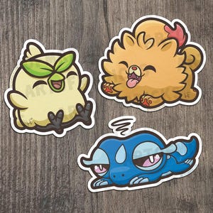 May include: Three cartoon stickers featuring cute animal characters. One is a yellow bird-like creature with green leaves, another is a fluffy orange dog with a pink bow, and the last is a blue creature with pink accents.