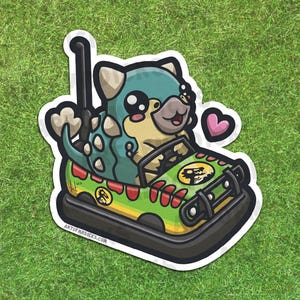 Puede incluir: Una pegatina con un híbrido de dinosaurio y perro de dibujos animados conduciendo un coche de choque. El coche es verde y amarillo con un logotipo de Jurassic Park. El dinosaurio-perro es verde azulado y marrón claro con ojos grandes y un corazón rosa. El texto "ART OF MR DICKS.COM" es visible.